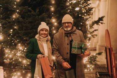 Happy senior couple enjoying outdoor christmas market at the evening, buying gifts and christmas tree. - HPIF32557
