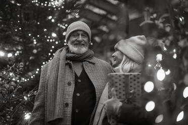 Black and white portrait of senior couple enjoying outdoor christmas market at the evening, buying gifts and christmas tree. - HPIF32558