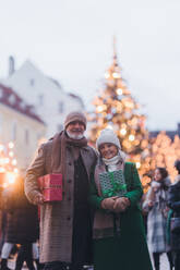 Happy senior couple enjoying outdoor christmas market in the city, buying gifts. Christmas square with a big Christmas tree in the center. - HPIF32560