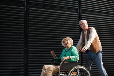 Porträt eines älteren Paares mit Frau im Rollstuhl. Älterer Mann schiebt seine schöne Frau im Rollstuhl. Konzept der chronischen Krankheit bei einem älteren Paar. Porträt mit Kopierraum auf schwarzem Hintergrund. Ältere Geschwister auf einem Sonntagsspaziergang in der Stadt. - HPIF32567