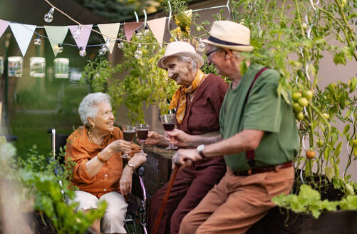 Porträt von älteren Freunden, die sich nach harter Arbeit im städtischen Garten in der Stadt bei einem Glas Wein entspannen. Rentner verbringen ihre Zeit gemeinsam bei der Gartenarbeit im Gemeinschaftsgarten in ihrer Wohnanlage. - HPIF32595