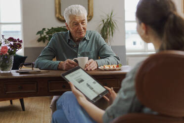 Caregiver reading online newspaper in digital tablet during taking care of senior man in his home. - HPIF32612