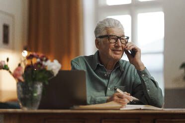 Senior man calling and writing some notes in his apartment. - HPIF32615