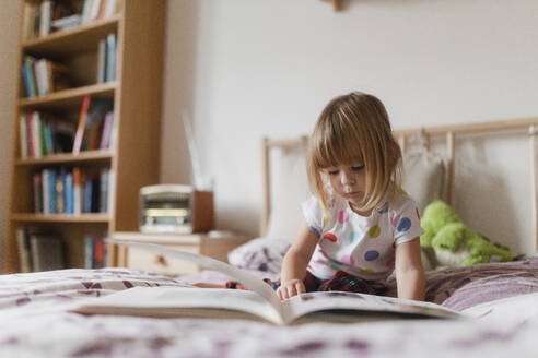 Cute little girl with bangs reading book lying on bed in her child's room. - HPIF32704