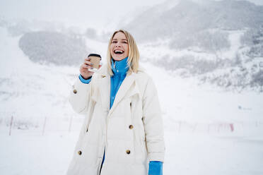 Glückliche blonde Frau hält Kaffeetasse und lacht auf schneebedecktem Berg - MDOF01760