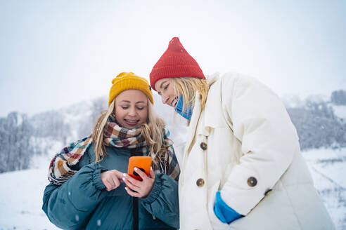 Lächelnde Frau benutzt ihr Smartphone und zeigt es einem Freund auf einem schneebedeckten Berg - MDOF01769