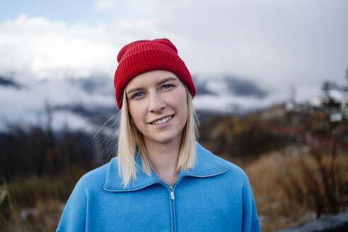 Lächelnde Frau mit roter Strickmütze unter winterlichem Himmel stehend - MDOF01772