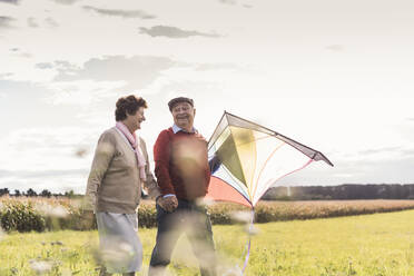Glückliches älteres Paar, das sich an den Händen hält und mit einem Drachen auf einem Feld spazieren geht - UUF30715