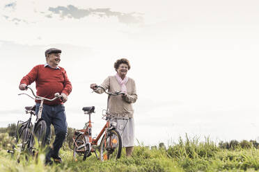 Happy senior couple walking with bicycles under sky - UUF30721