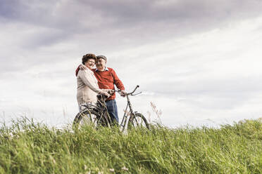 Happy senior couple standing with bicycle at field under cloudy sky - UUF30723