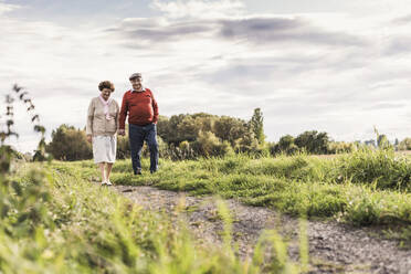 Senior couple holding hands and walking on footpath under cloudy sky - UUF30729