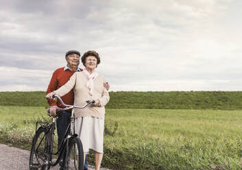 Smiling senior couple standing with bicycle on footpath at field - UUF30734