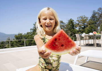 Lächelndes Mädchen sitzt auf einem Liegestuhl und hält eine Scheibe Wassermelone in der Hand - NJAF00694