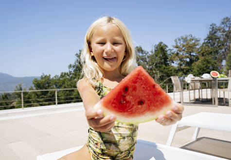 Lächelndes Mädchen sitzt auf einem Liegestuhl und hält eine Scheibe Wassermelone in der Hand - NJAF00694