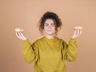 Smiling woman with eyes closed holding doughnuts against peach background - MMPF01072