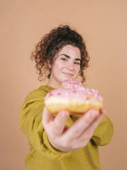 Young woman showing doughnut against peach background - MMPF01073