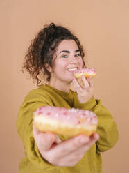 Smiling young woman showing and holding doughnuts against peach background - MMPF01074