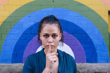 Woman with finger on lips in front of rainbow wall - ASGF04787