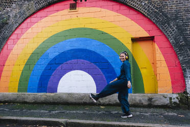 Glückliche Frau mit kabellosen Kopfhörern, die vor einer Regenbogenwand genießt - ASGF04796