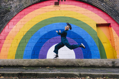 Happy young woman wearing wireless headphones and dancing in front of rainbow wall - ASGF04800