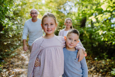 Portrait of little children, siblings, hugging in a forest, their parent walking in the background. - HPIF32769
