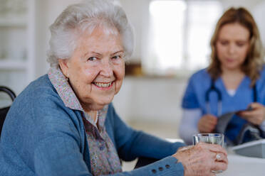 Portrait of senior woman with glass of water, the nurse assistant sitting in background. - HPIF32802