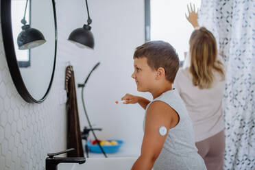 The diabetic boy brushing his teeth in the morning, preparing for the day, while wearing a continuous glucose monitoring sensor on his arm. CGM device making life of school boy easier, helping manage his illness and focus on other activities. - HPIF32891