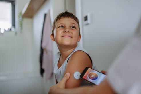 Junge mit Diabetes, der seinen Blutzuckerspiegel zu Hause mit einem kontinuierlichen Glukosemessgerät kontrolliert. Die Mutter des Jungen verbindet sein CGM mit einem Smartphone, um seinen Blutzuckerspiegel in Echtzeit zu überwachen. - HPIF32903