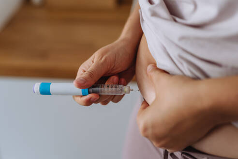 Close up of needle injecting insulin in abdomen in bathroom. Woman with type 1 diabetes taking insuling with syringe needle. - HPIF32922