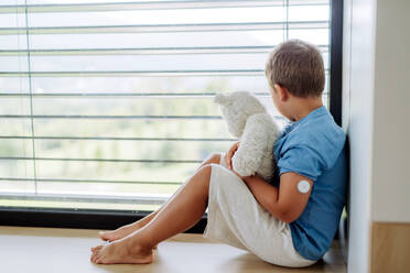 Diabetic boy with a continuous glucose monitor sitting by the window, holding his stuffed teddy bear and looking outside. Children with diabetes feeling different or isolated from peers. - HPIF32925