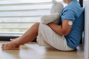 Diabetic boy with a continuous glucose monitor sitting by the window, holding his stuffed teddy bear and looking outside. Children with diabetes feeling different or isolated from peers. - HPIF32926