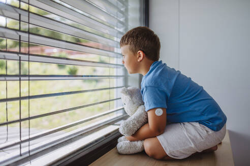 Diabetikerjunge mit kontinuierlichem Blutzuckermessgerät, der am Fenster sitzt, seinen Plüschteddy hält und nach draußen schaut. Kinder mit Diabetes fühlen sich anders oder von Gleichaltrigen isoliert. - HPIF32928