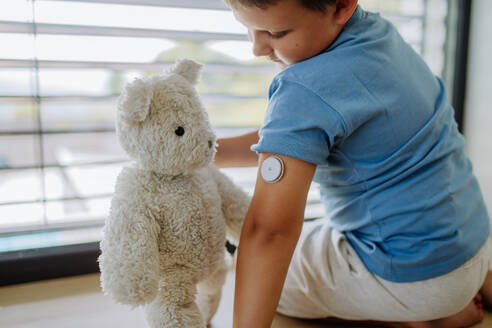 Diabetikerjunge mit einem kontinuierlichen Blutzuckermessgerät, der am Fenster sitzt und seinen ausgestopften Teddybär-Sensor auf dem Arm zeigt. Kinder mit Diabetes fühlen sich anders oder von Gleichaltrigen isoliert. - HPIF32930