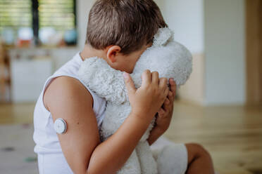 Diabetikerjunge mit kontinuierlichem Blutzuckermessgerät, der zu Hause sitzt und seinen Plüschteddy umarmt. Kinder mit Diabetes fühlen sich anders oder von Gleichaltrigen isoliert. - HPIF32933