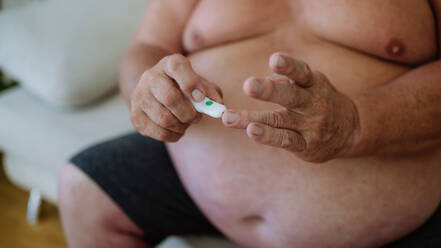 Übergewichtiger Mann, der seinen Blutzuckerspiegel zu Hause mit einem Blutzuckermessgerät mit Fingerstich kontrolliert. Der Diabetiker, der eine Blutprobe aus dem Finger entnimmt. - HPIF32958