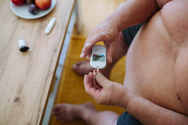 Übergewichtiger Mann, der seinen Blutzuckerspiegel zu Hause mit einem Blutzuckermessgerät mit Fingerstich kontrolliert. Der Diabetiker, der eine Blutprobe aus dem Finger entnimmt. - HPIF32960