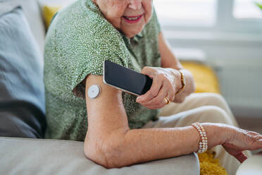 Nahaufnahme einer älteren Diabetikerin, die ihren Blutzuckerspiegel zu Hause mit einem kontinuierlichen Glukosemessgerät überprüft. Eine ältere Frau verbindet ihr CGM mit einem Smartphone, um ihren Blutzuckerspiegel in Echtzeit zu sehen. - HPIF33159