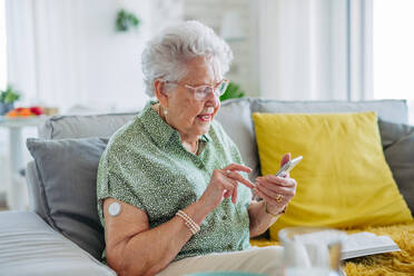 Ältere Diabetikerin mit kontinuierlichem Blutzuckermessgerät zur Kontrolle des Blutzuckerspiegels zu Hause. Das CGM-Gerät erleichtert das Leben einer älteren Frau und hilft ihr, mit ihrer Krankheit umzugehen und sich auf andere Aktivitäten zu konzentrieren. Ältere Diabetikerin, die ihre Blutzuckerdaten auf dem Smartphone überprüft. - HPIF33164