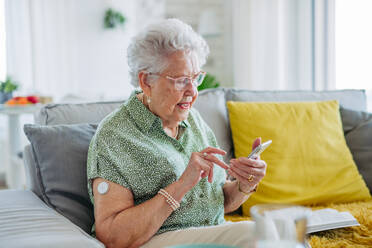 Diabetic senior patient using continuous glucose monitor to check blood sugar level at home. CGM device making life of elderly woman easier, helping manage her illness and focus on other activities. Senior diabetic woman checking her glucose data on smartphone. - HPIF33164