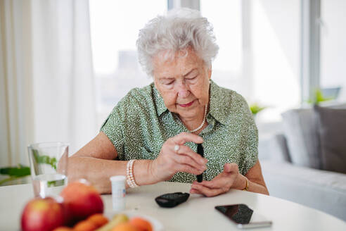 Diabetische ältere Patientin, die ihren Blutzuckerspiegel mit einem Glukosemessgerät mit dem Finger prüft. Porträt einer älteren Frau mit Typ-1-Diabetes, die zu Hause ein Blutzuckermessgerät benutzt. - HPIF33184