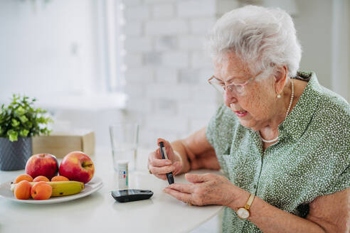 Porträt einer älteren Diabetikerin, die ihren Blutzuckerspiegel mit einem Glukosemessgerät mit Fingerstich überprüft. Porträt einer älteren Frau mit Typ-1-Diabetes, die zu Hause ein Blutzuckermessgerät benutzt. - HPIF33188