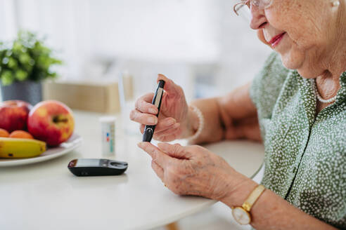 Diabetic senior patient checking her blood sugar level with fingerstick testing glucose meter. Portrait of senior woman with type 1 diabetes using blood glucose monitor at home. - HPIF33189