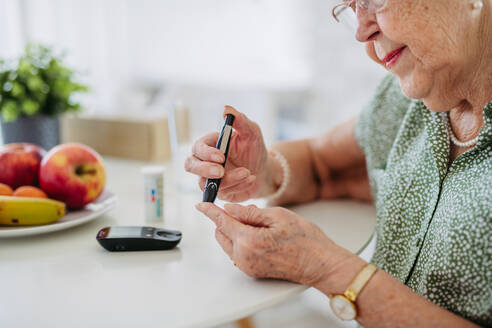 Diabetische ältere Patientin, die ihren Blutzuckerspiegel mit einem Glukosemessgerät mit dem Finger prüft. Porträt einer älteren Frau mit Typ-1-Diabetes, die zu Hause ein Blutzuckermessgerät benutzt. - HPIF33189