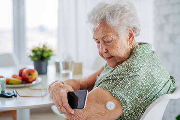 Diabetic senior patient checking blood glucose level at home using continuous glucose monitor. Elderly woman connecting her CGM with smarphone to see her blood sugar levels in real time. - HPIF33190