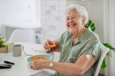 Ältere Diabetikerin mit kontinuierlichem Glukosemessgerät zur Kontrolle des Blutzuckerspiegels zu Hause. Ältere Frau isst Obst, um ihren Blutzuckerspiegel auf ein normales Niveau zu bringen. CGM-Gerät, das das Leben einer älteren Frau erleichtert und ihr hilft, ihre Krankheit zu bewältigen und sich auf andere Aktivitäten zu konzentrieren. - HPIF33195