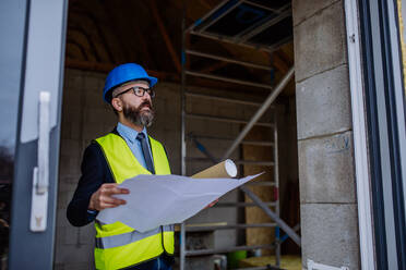 Mature businessman controlling blueprints from an unfinished house. - HPIF33302