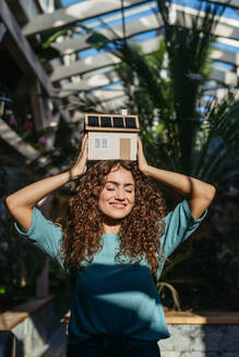 Junge Frau in einem botanischen Garten, die ein Modell eines Hauses mit Sonnenkollektoren hält, Konzept der grünen Energie. - HPIF33322