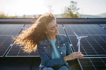 Portrait of young woman, holding model of a wind turbine on roof with solar panels. - HPIF33345
