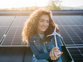 Portrait of young woman, holding model of a wind turbine on roof with solar panels. - HPIF33346