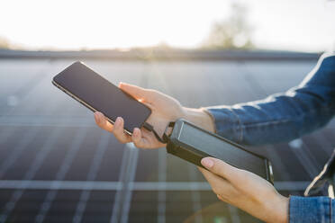 Close up of a woman charging her phone with solar charger on the roof with photovoltaics panels. - HPIF33352